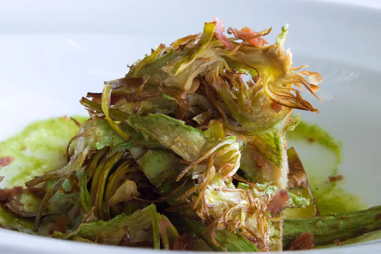 Fried artichoke slices with crispy jamon bits in a white bowl with green sauce