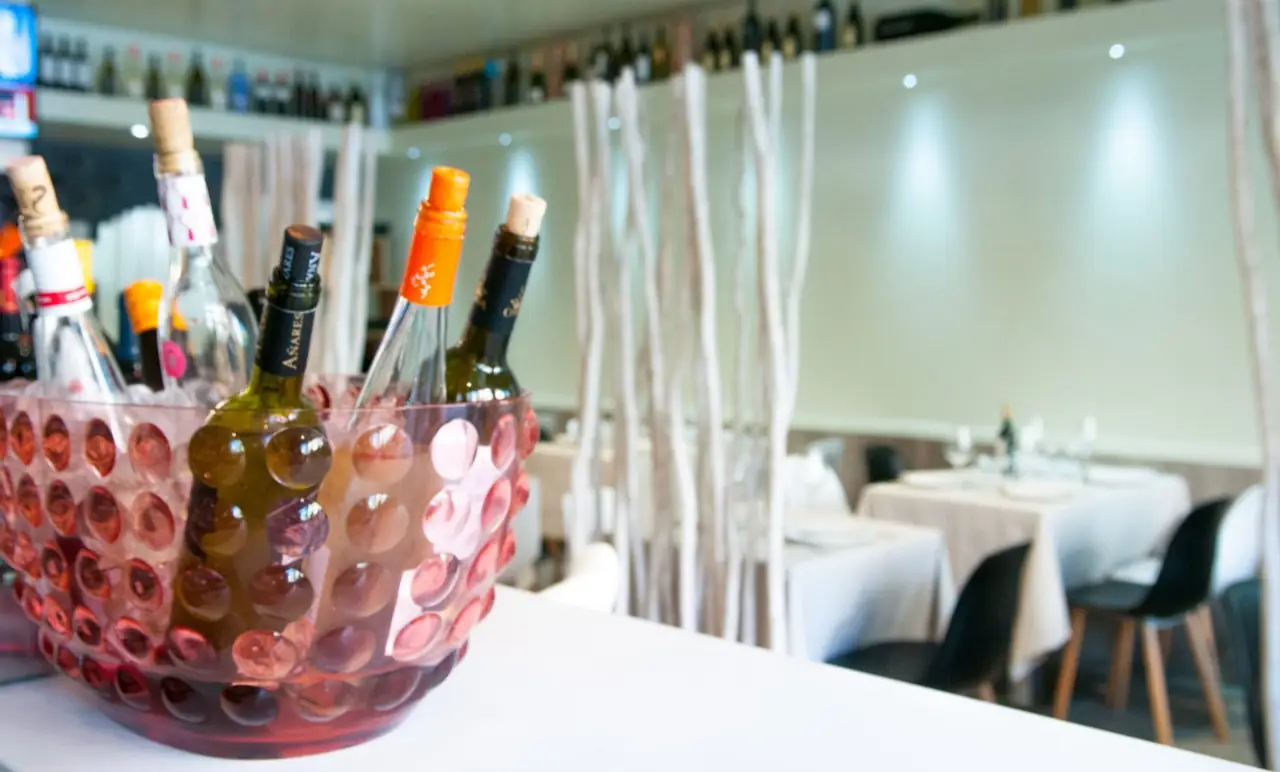 Dining room interior with white-linen tables behind a dimpled ice bucket holding chilling wine bottles, shelves of wine on the back wall