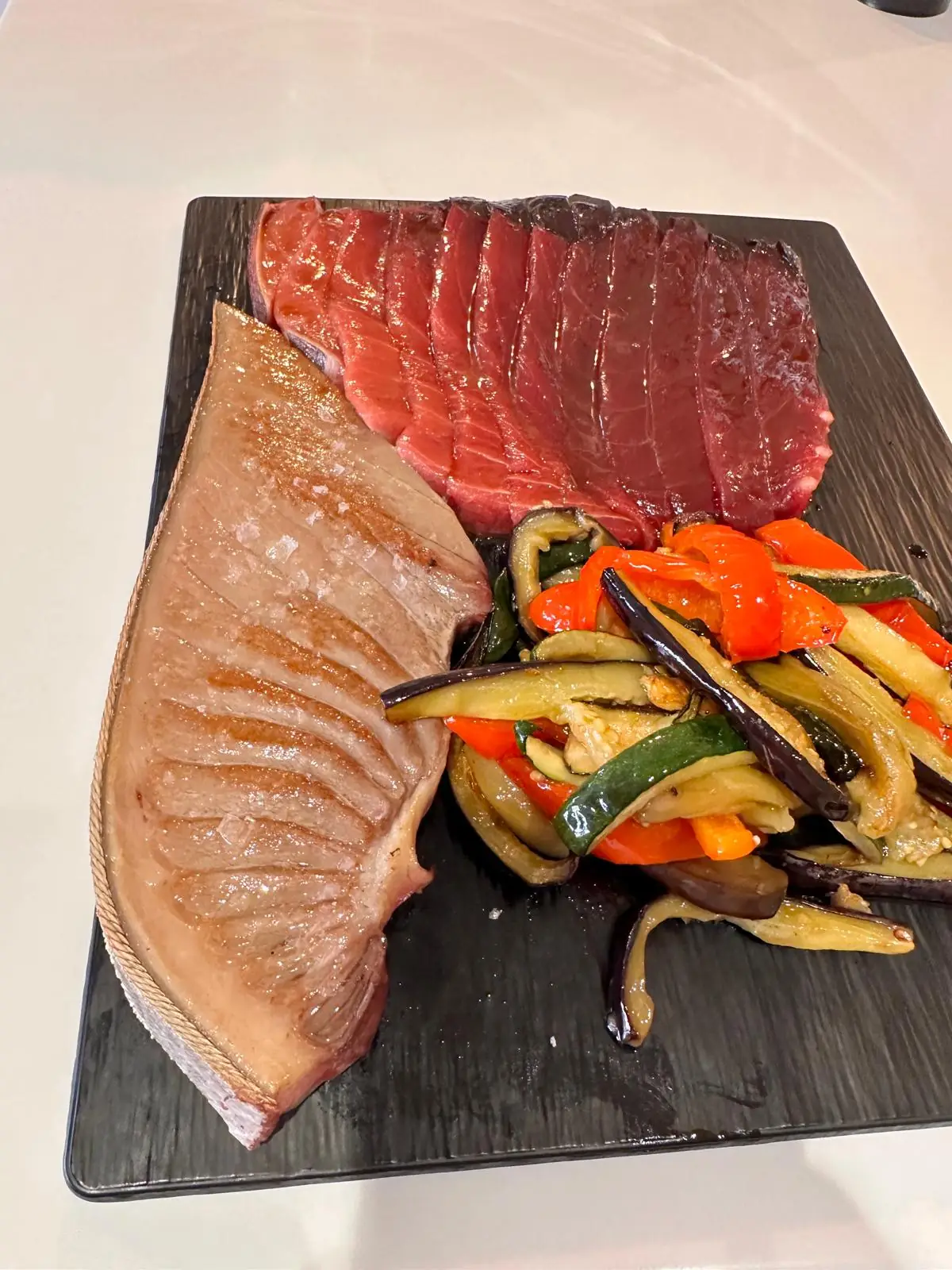Two thick slices of seared red tuna with sauteed courgette, aubergine and red pepper on a black slate board