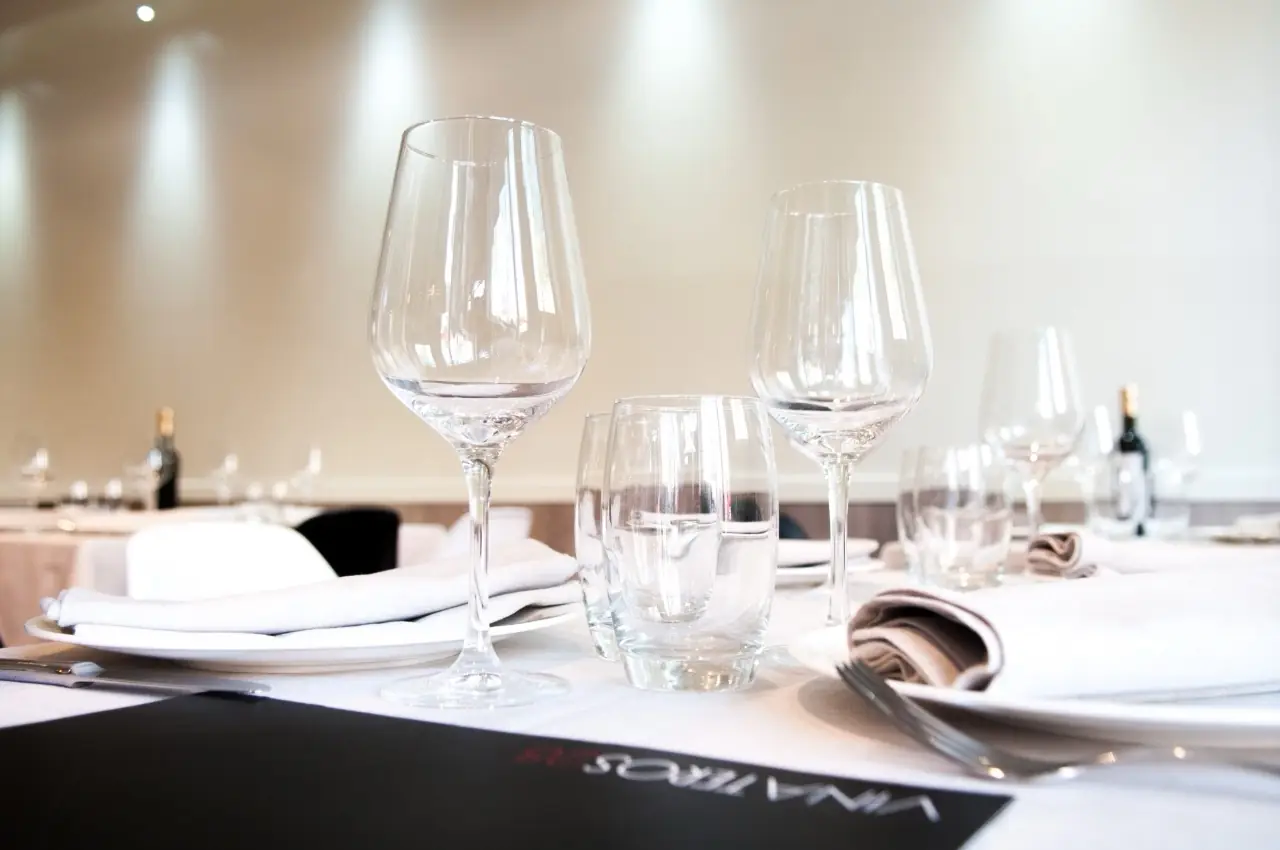 Table setting with wine glasses, water tumblers, folded napkins and a black Vinateros-branded menu card, dining room behind