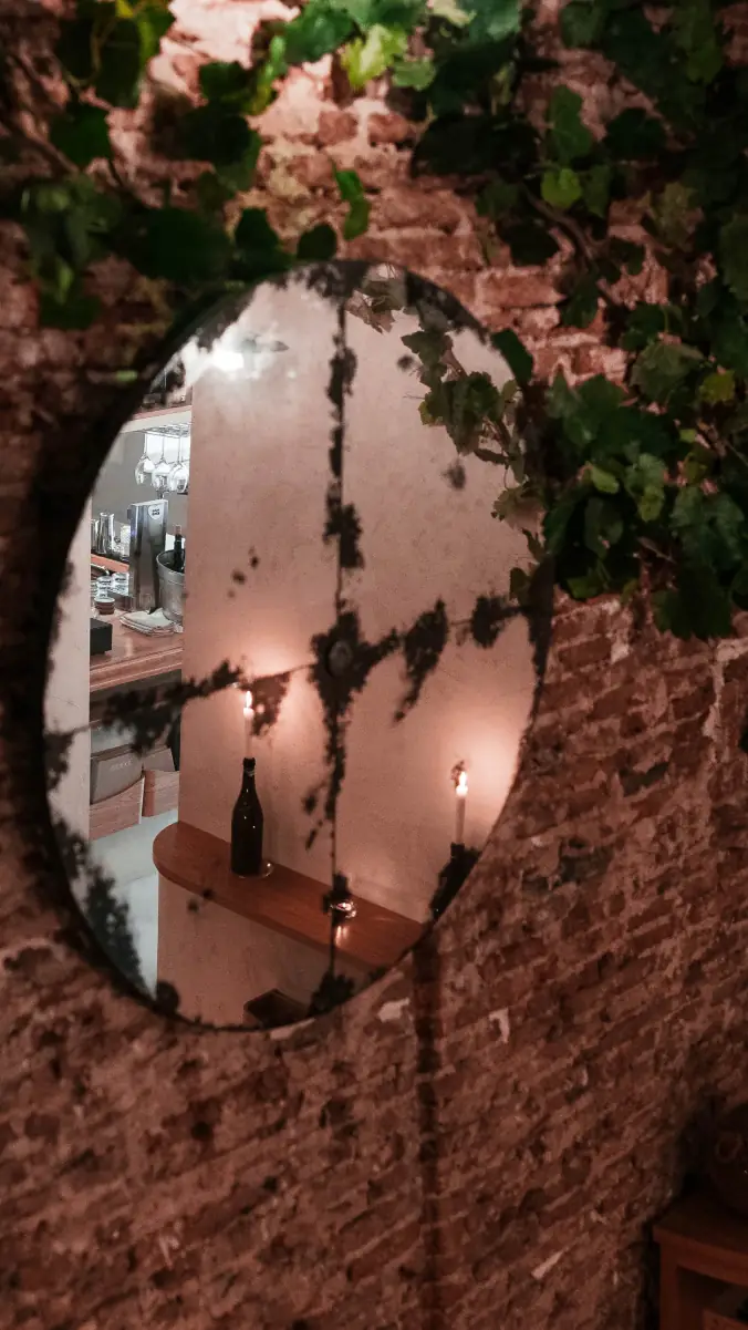 Round mirror mounted on an exposed brick wall framed by trailing vine leaves, reflecting a candle-lit bar with a wine bottle on a wooden shelf