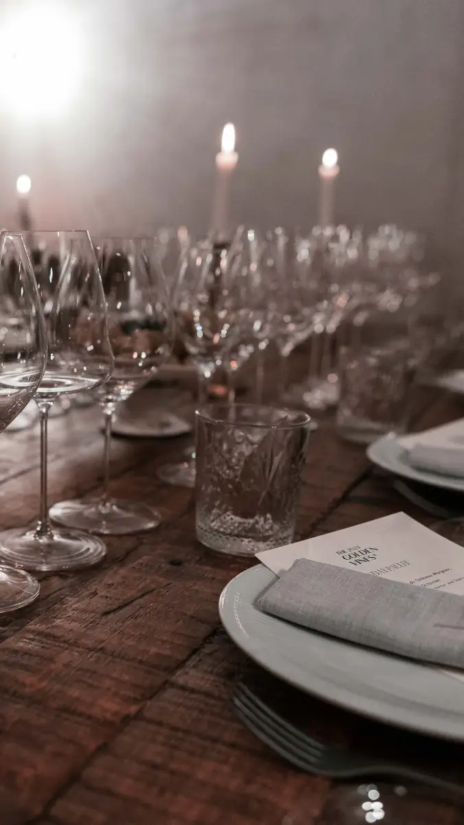 Rustic wooden table set for a tasting with rows of wine glasses, a cut-crystal tumbler, lit taper candles, plates and a folded napkin over a printed menu