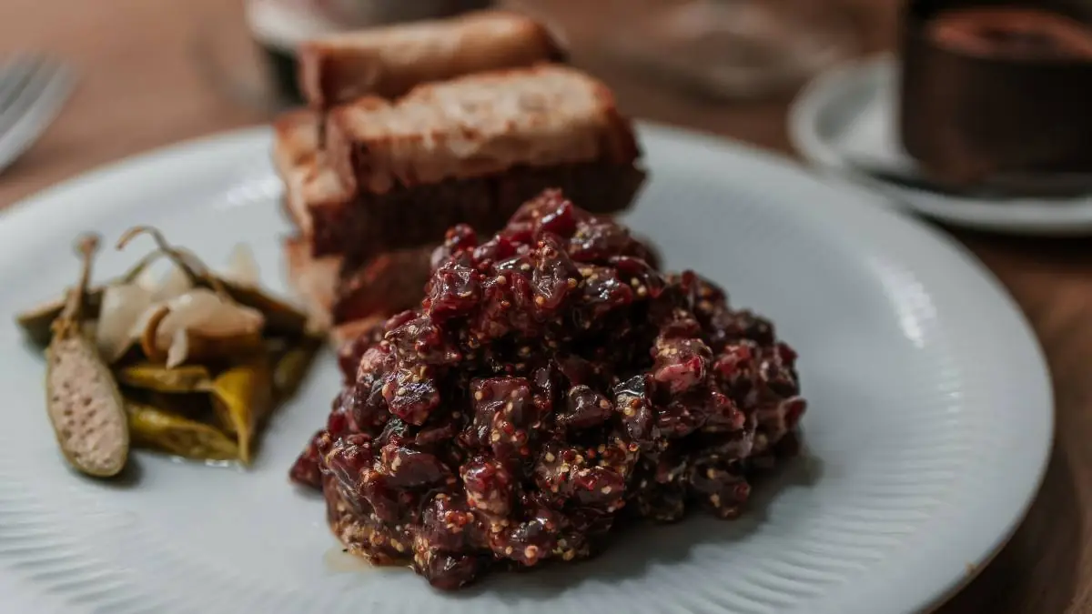 Mound of steak tartare studded with mustard seeds on a white plate with pickled cornichons, peppers and slices of toasted bread