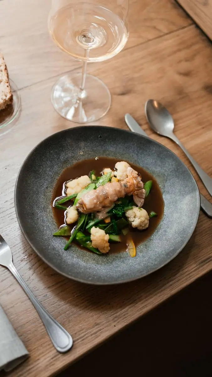 Langoustine tail with cauliflower florets, snap peas and greens in a brown broth in a grey bowl, with a glass of white wine on a wooden table