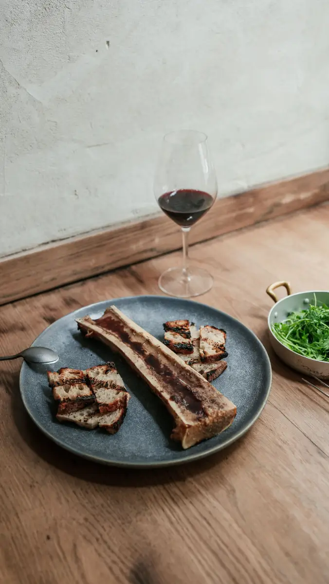 Split roasted bone marrow with grilled bread slices on a grey plate, a side dish of arugula and a glass of red wine on a wooden table