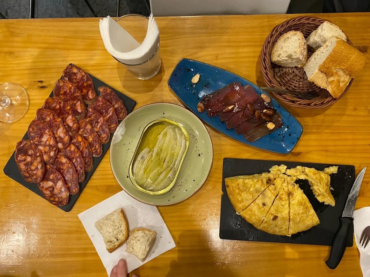 A wooden table with sliced cured meats, tinned fish, a tortilla wedge and bread