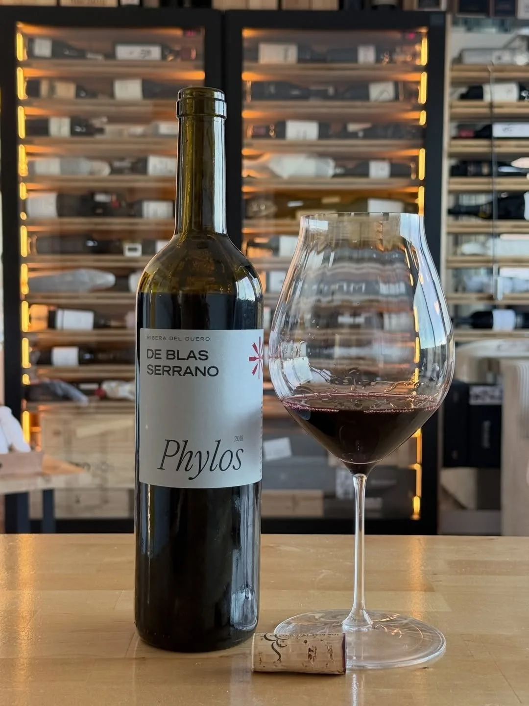A bottle of red wine and a full glass on a wooden counter in front of wine racks