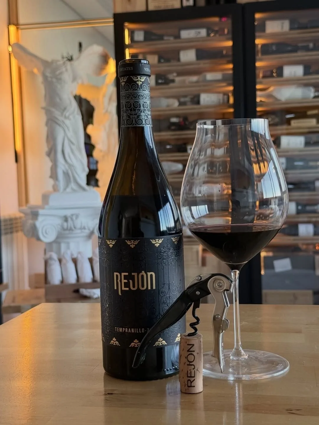 A bottle of red wine and a glass on a wooden counter in front of wine racks