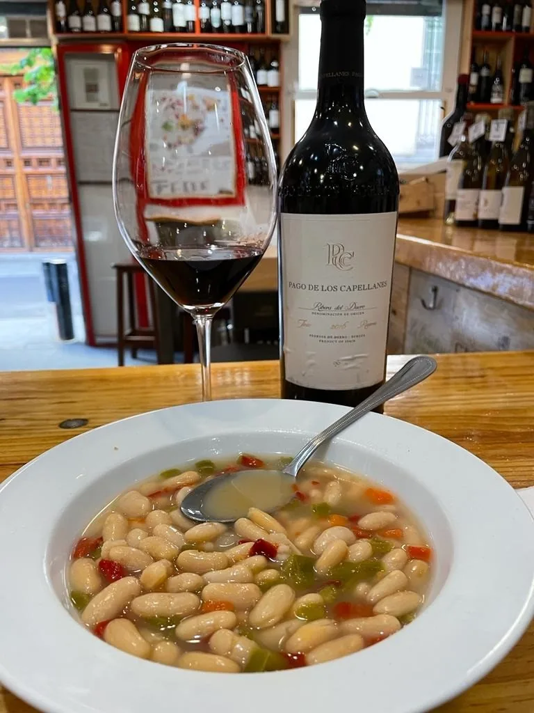 A bowl of white bean stew beside a glass and bottle of red wine