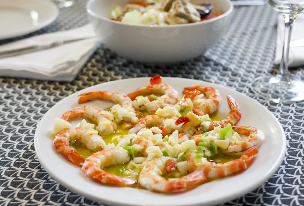 White plate of whole cooked shrimp with tails on, topped with finely chopped mixed vegetables in olive oil