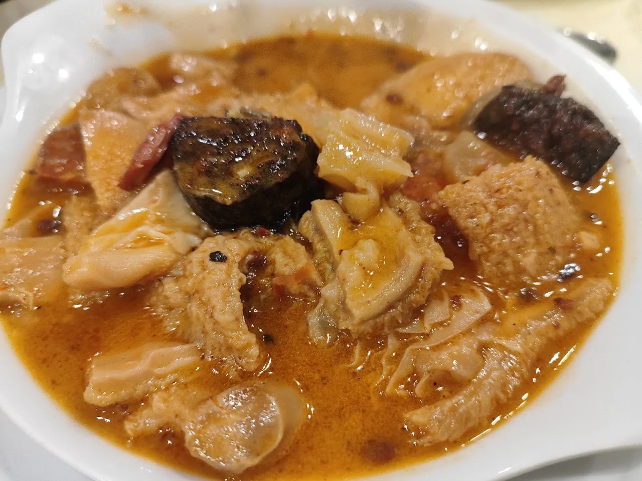 Close-up bowl of a brown stew with tender meat pieces, chorizo and morcilla slices in a thick broth
