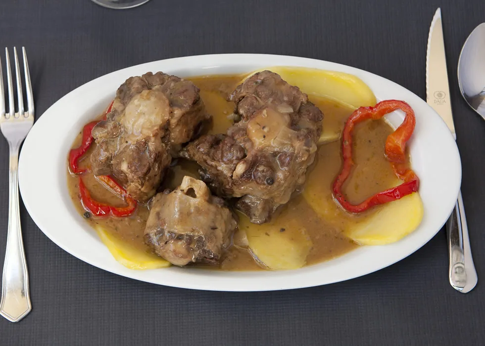 Overhead shot of a white oval plate with braised meat chunks in a brown sauce, sliced potatoes and strips of red pepper