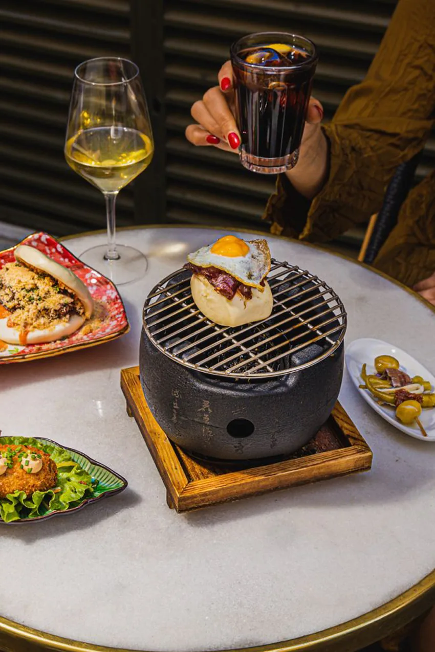 A round marble table spread with a filled bao, a croqueta on lettuce, a plate of gilda skewers with olives and anchovies, and a bao topped with a fried quail egg, alongside a glass of white wine and a glass of vermouth