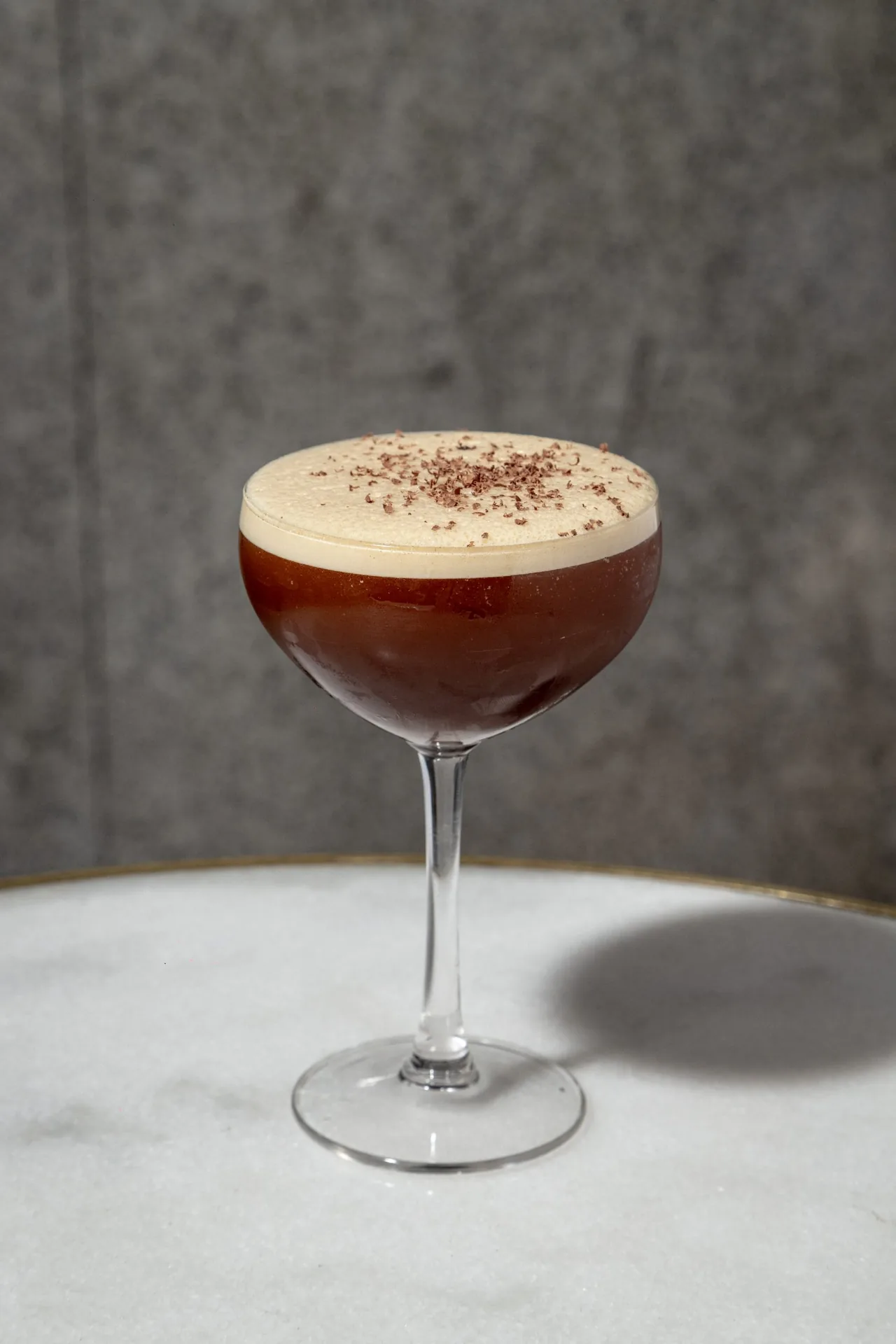 An espresso martini in a coupe with a thick pale foam head dusted with grated chocolate, on a marble table against a grey plaster wall