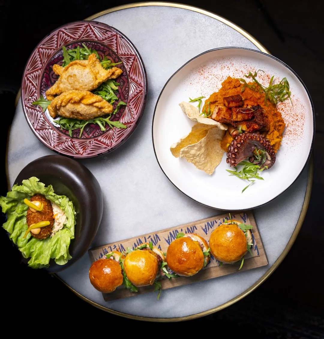 Overhead spread of fried empanadas, a plate of octopus with sweet potato puree and crackling, a lettuce-wrapped croqueta, and four glazed brioche sliders on a wooden board