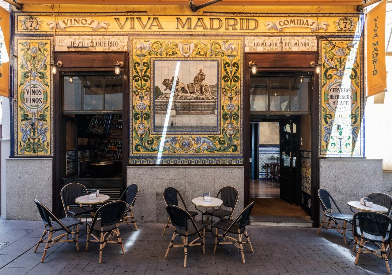 A colourful tiled frontage with lettering above a doorway and windows, and small cafe tables and chairs outside.
