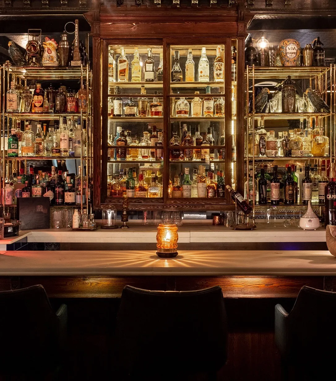 A back bar with shelves of bottles lit by a candle on a dark counter.