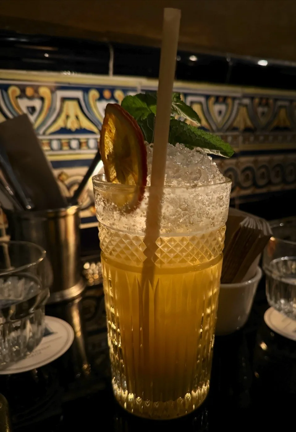 A tall cocktail with a straw and citrus slice against a patterned tiled wall.