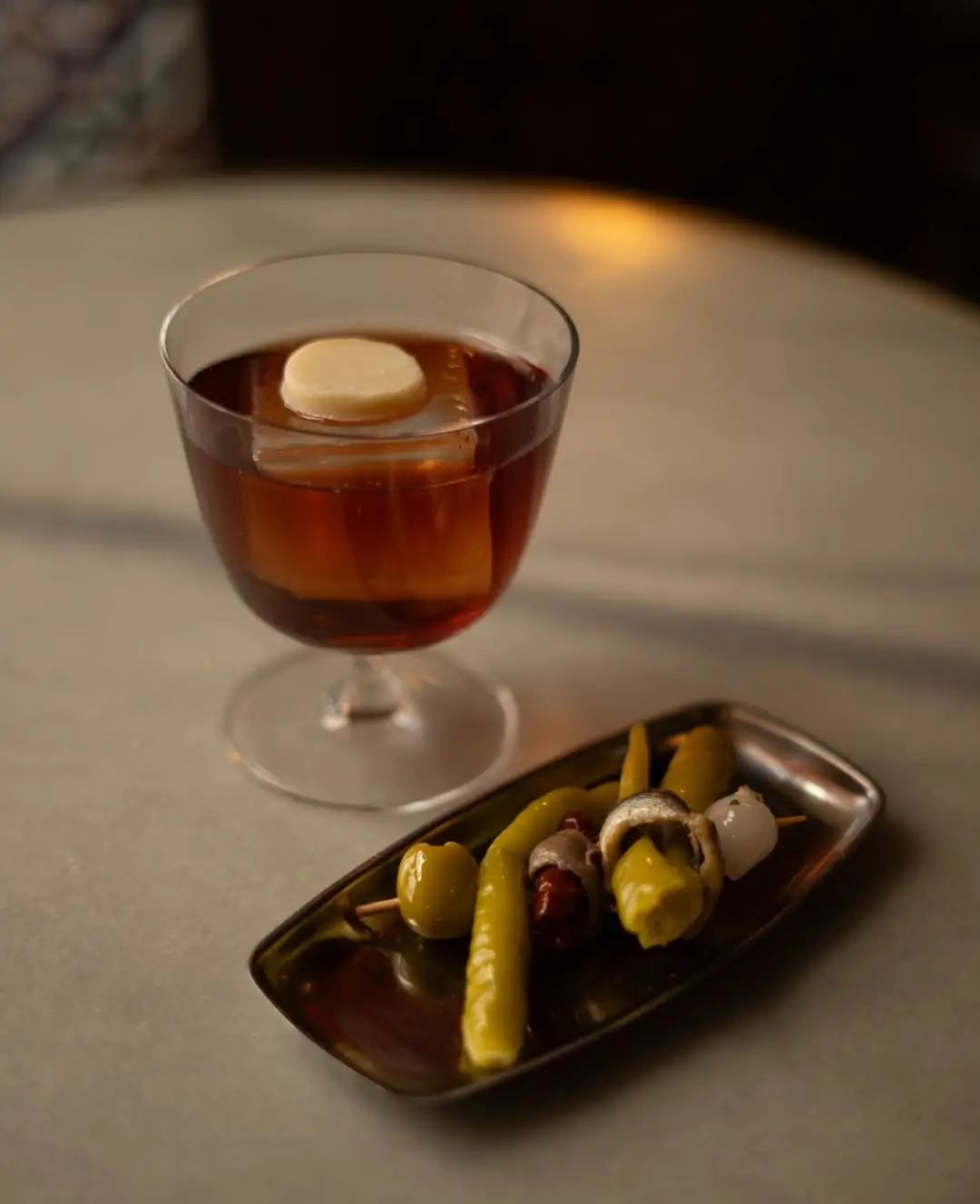 Amber vermouth in a glass with an ice cube beside a metal tray of olive and pickle skewers