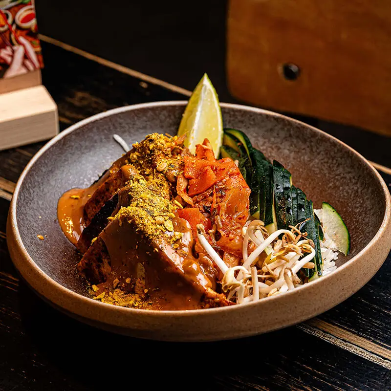 Bowl with a marinated tofu steak, kimchi, cucumber ribbons, bean sprouts, rice, and a lime wedge, topped with crushed pistachio and chili at Vrutal