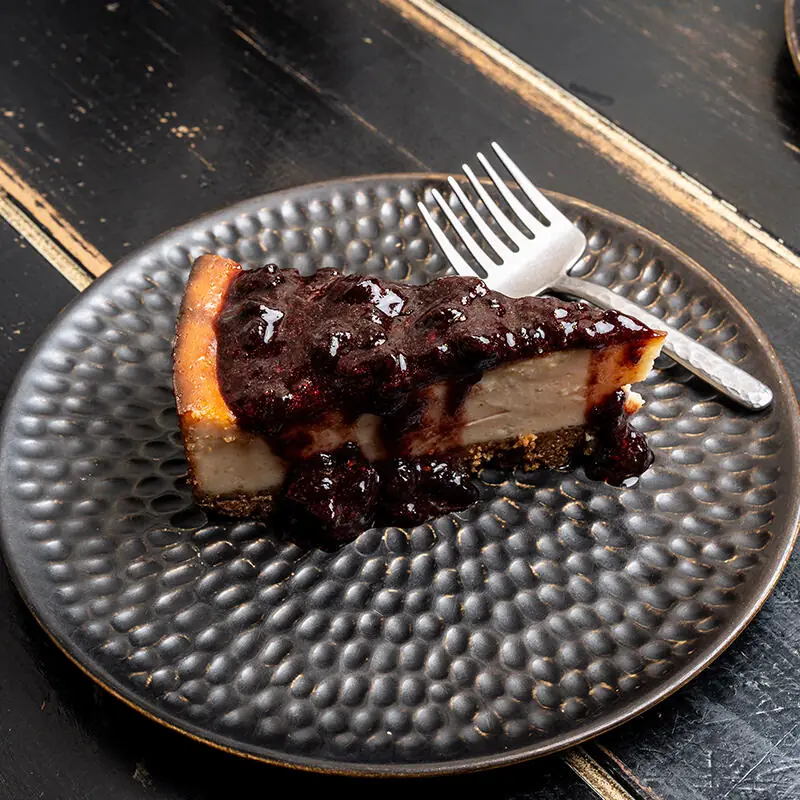 Slice of cheesecake with a berry compote topping and graham crust, served on a dark textured plate with a fork at Vrutal