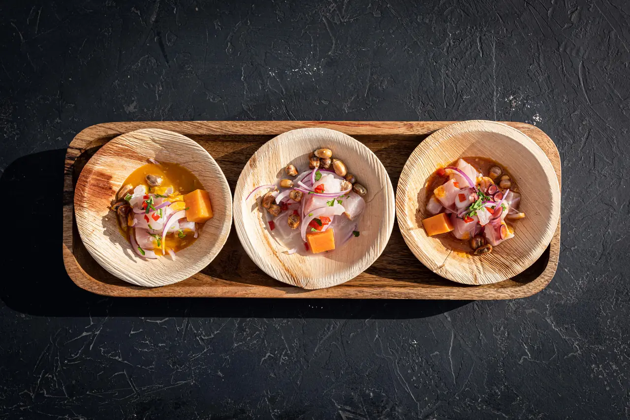 Three ceviche tostadas served in a wooden tray, with orange sea bass in citrus leche de tigre, red onion, and Peruvian corn kernels, Warike Project
