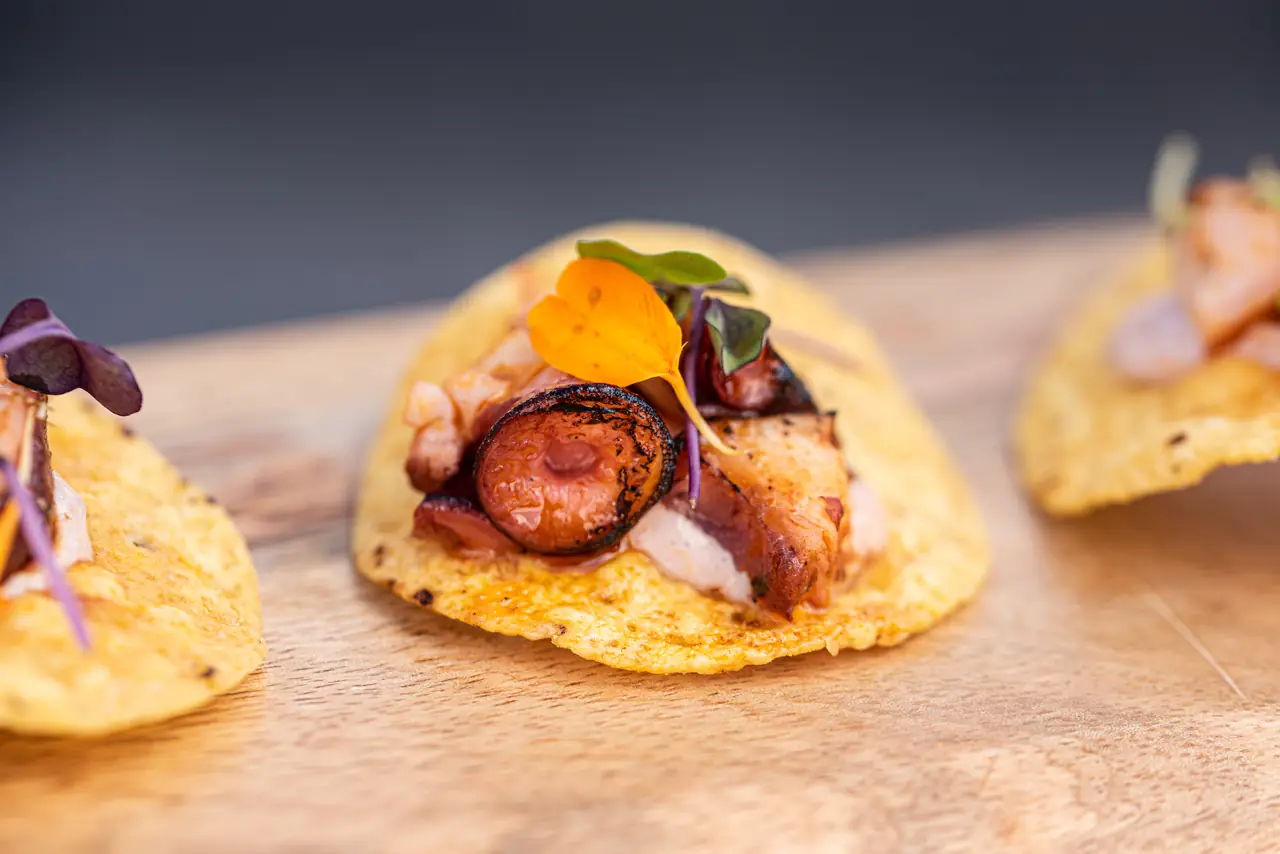 Crispy corn tostada topped with seared octopus, shrimp, and a small edible flower on a wooden board, Warike Project