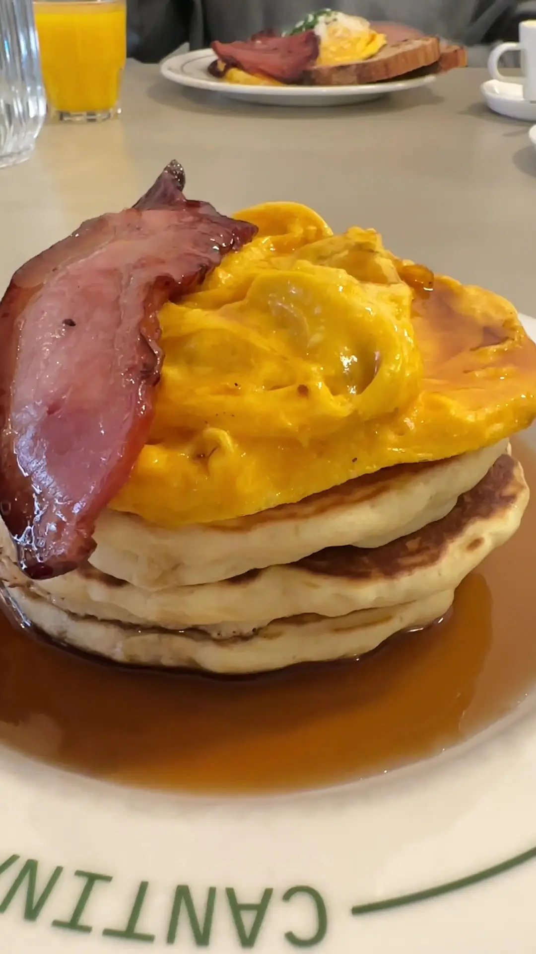A stack of pancakes topped with scrambled eggs and bacon in syrup, on a Watts Cantina plate