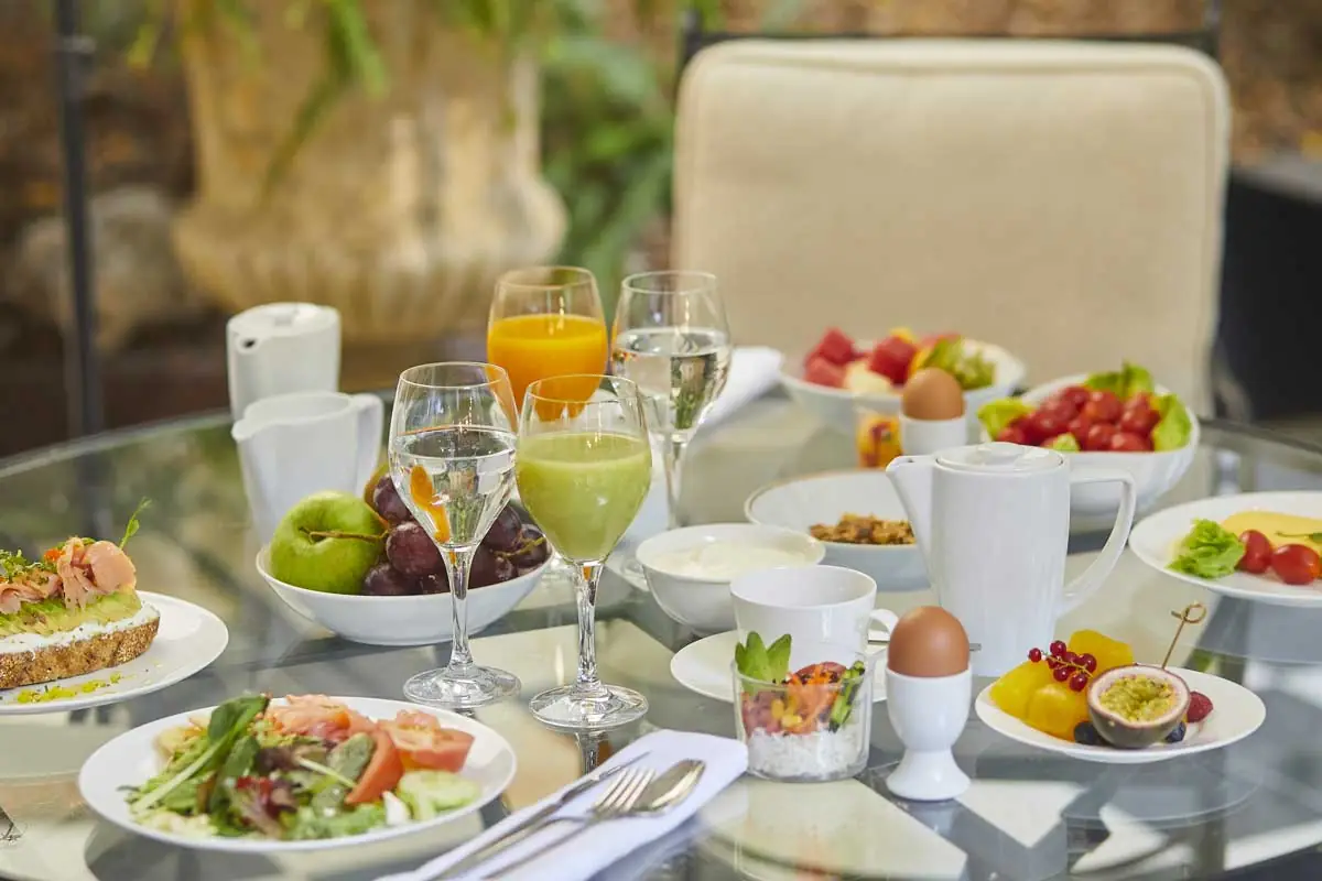 Breakfast spread on a glass terrace table with a smoked salmon tartine, mixed salad, fresh fruit, yogurt, juices, and a soft-boiled egg, at Winter Garden