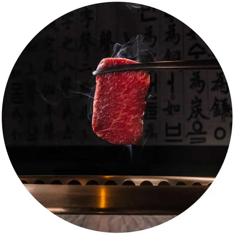 Slice of marbled beef held with chopsticks, smoke rising, over a round grill against a wall of Asian calligraphy
