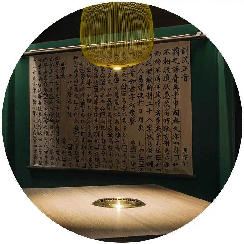 Dining nook with a hanging calligraphy scroll on a green wall, yellow pendant lamp, and a wooden table with an inset grill