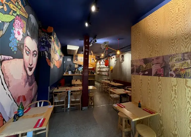 Narrow dining room with a large painted geisha mural on a navy wall, wooden tables and stools, and a plywood partition with sushi graphics
