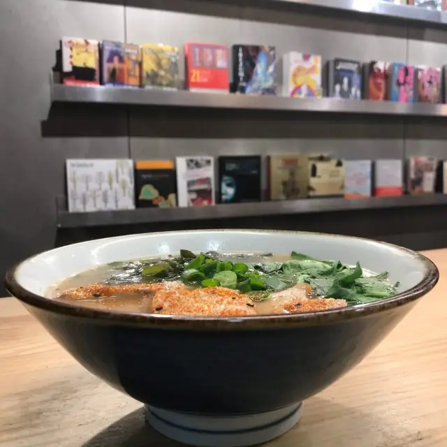 Bowl of ramen with broth, greens, spring onion and a chili-dusted topping on a wooden counter with a bookshelf behind