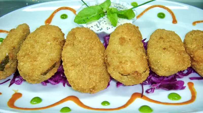 Row of breaded stuffed jalapeno croquettes on shredded red cabbage with a white dip and mint garnish on a white plate