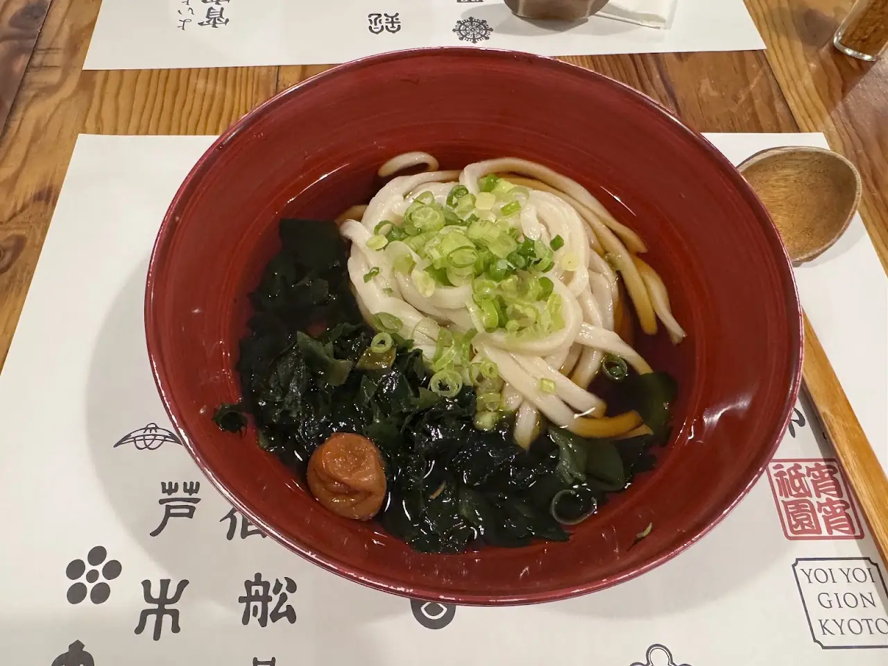 Bowl of udon noodles in broth with wakame seaweed and scallions in a red bowl, at Yoi Yoi Gion.