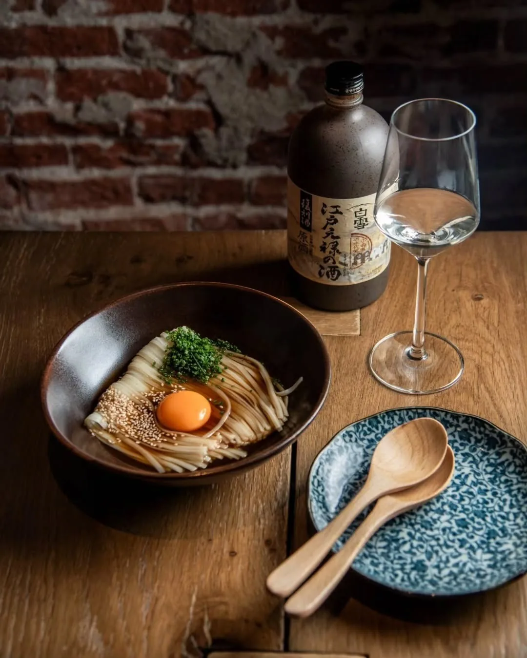 A dark bowl of noodles topped with a raw egg yolk beside a sake bottle and a glass