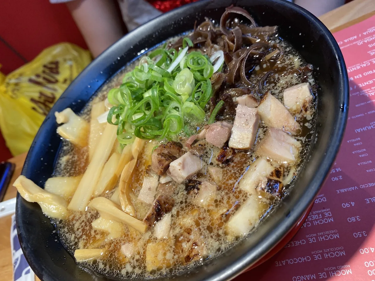 A black bowl of ramen in broth with chunks of pork, bamboo shoots, wood ear mushrooms and chopped spring onion