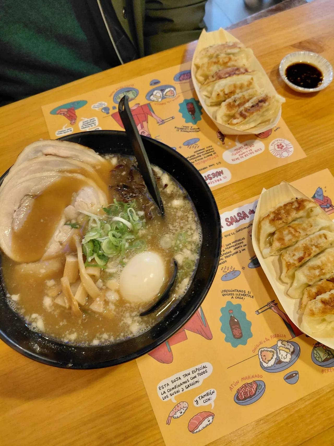 A black bowl of ramen with sliced pork, a soft-boiled egg and spring onion beside a tray of gyoza