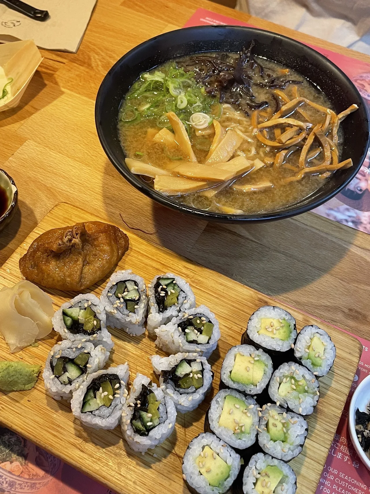 A bowl of ramen with bamboo shoots, wood ear mushrooms and orange strips on a wooden board beside maki rolls and an inari pouch