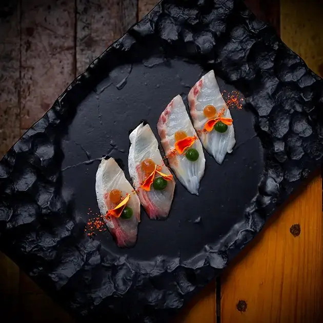 Four slices of pale white fish sashimi laid diagonally across a rough black slate, each dotted with a green pearl, an orange gel bead and a curl of orange petal, on a wooden table