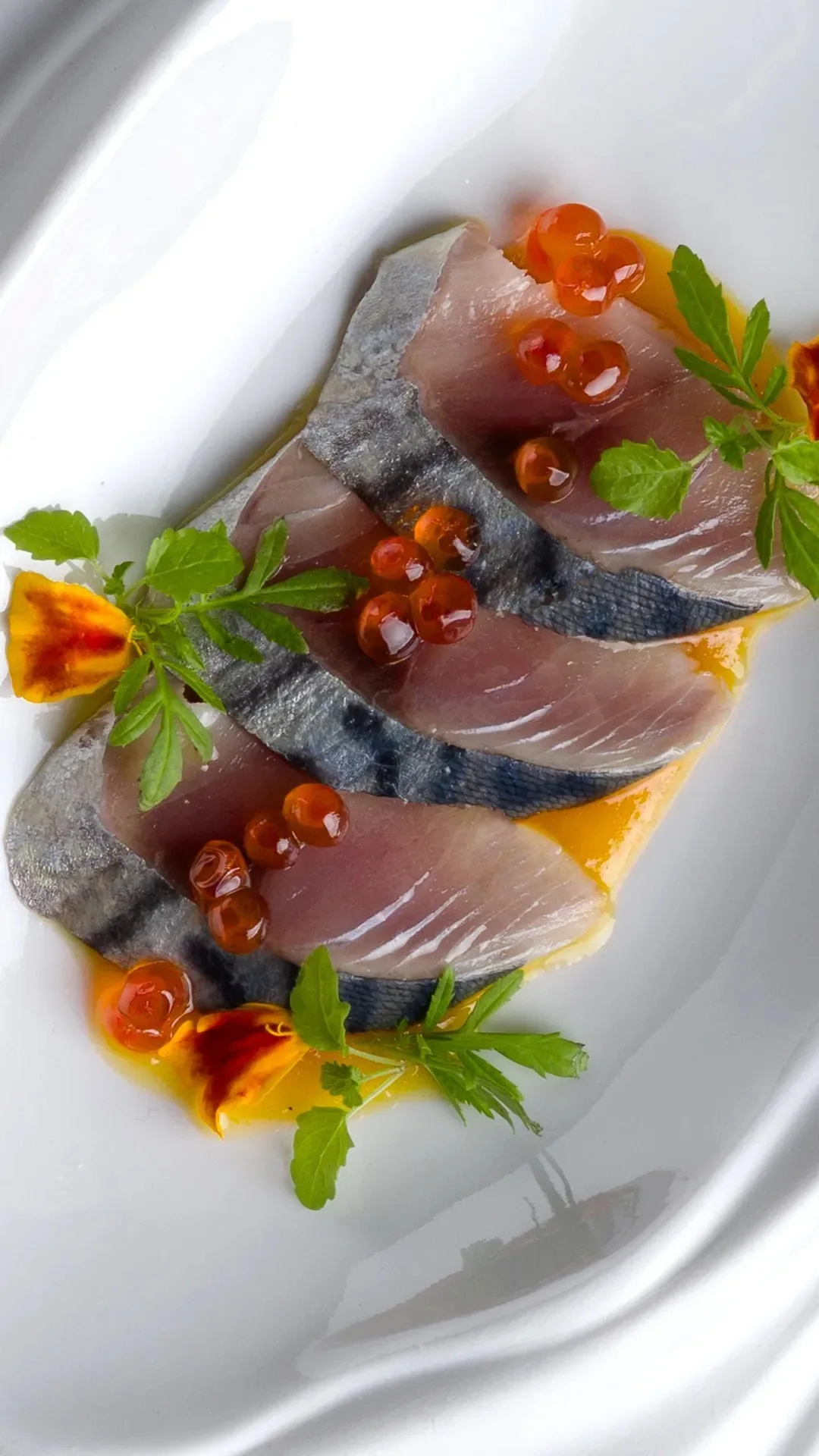 Thin slices of fish topped with red roe and small herbs on a white plate