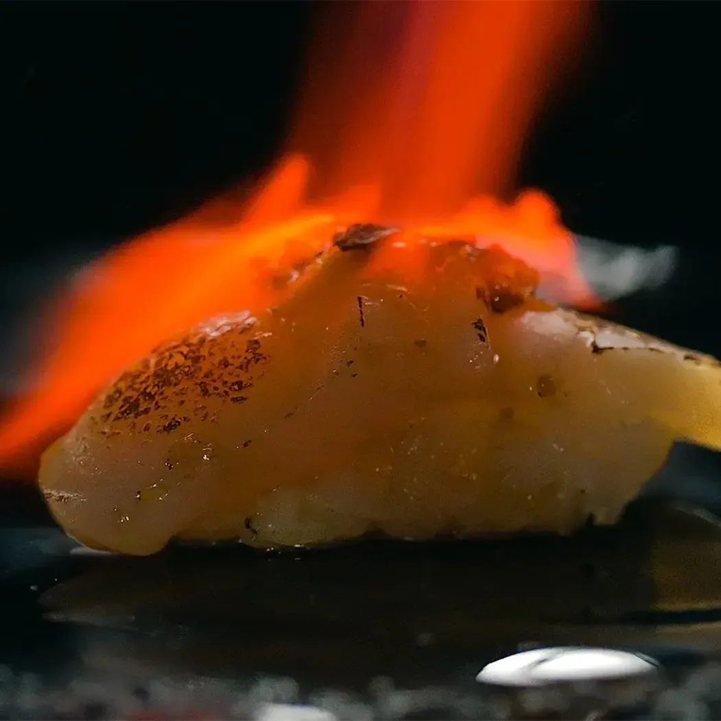 A torched piece of nigiri with flames above it on a dark plate