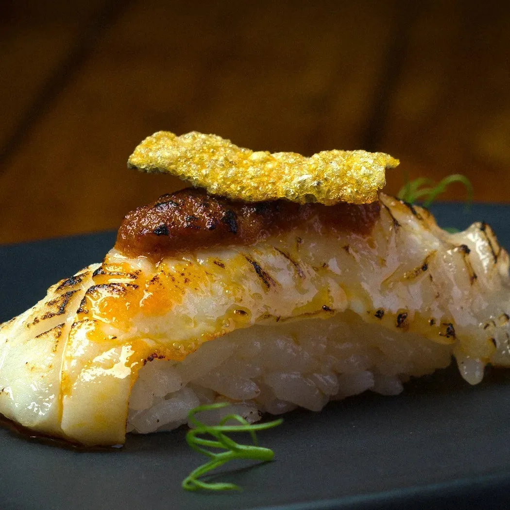 A piece of nigiri with seared white fish and a golden topping on a dark plate