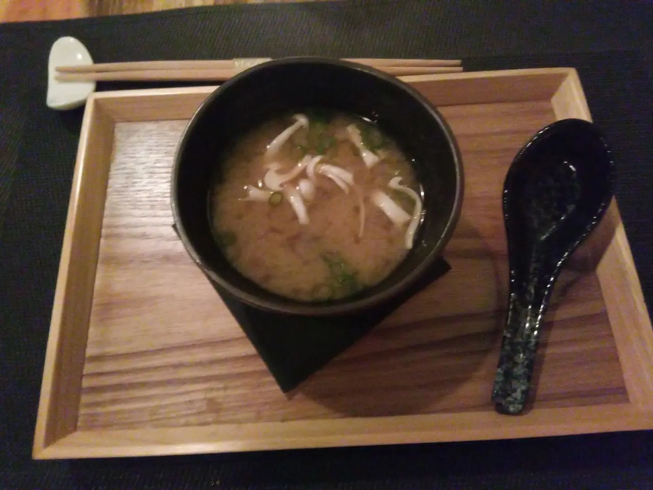 A bowl of miso soup with sliced mushrooms, served on a wooden tray with chopsticks and a black spoon.