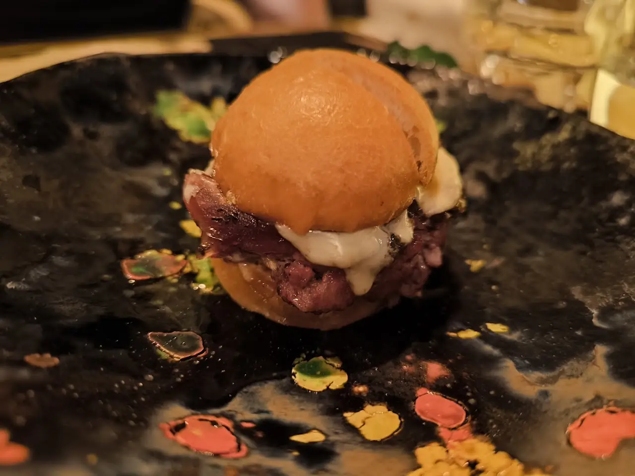 A small brioche slider filled with sliced meat and melted cheese on a black splattered plate.