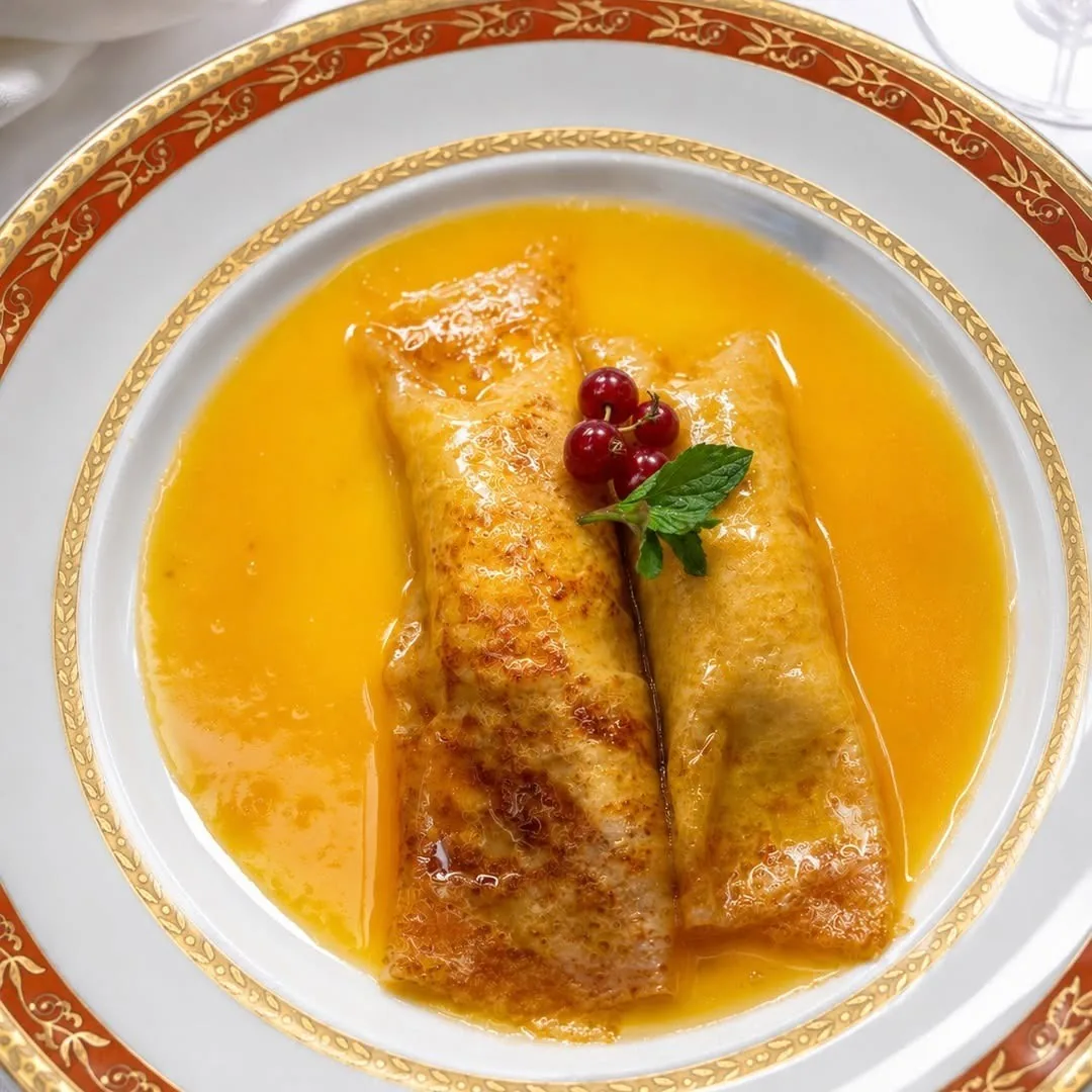 Two rolled golden crepes in orange sauce with a red berry and mint leaf on a plate with a red and gold rim