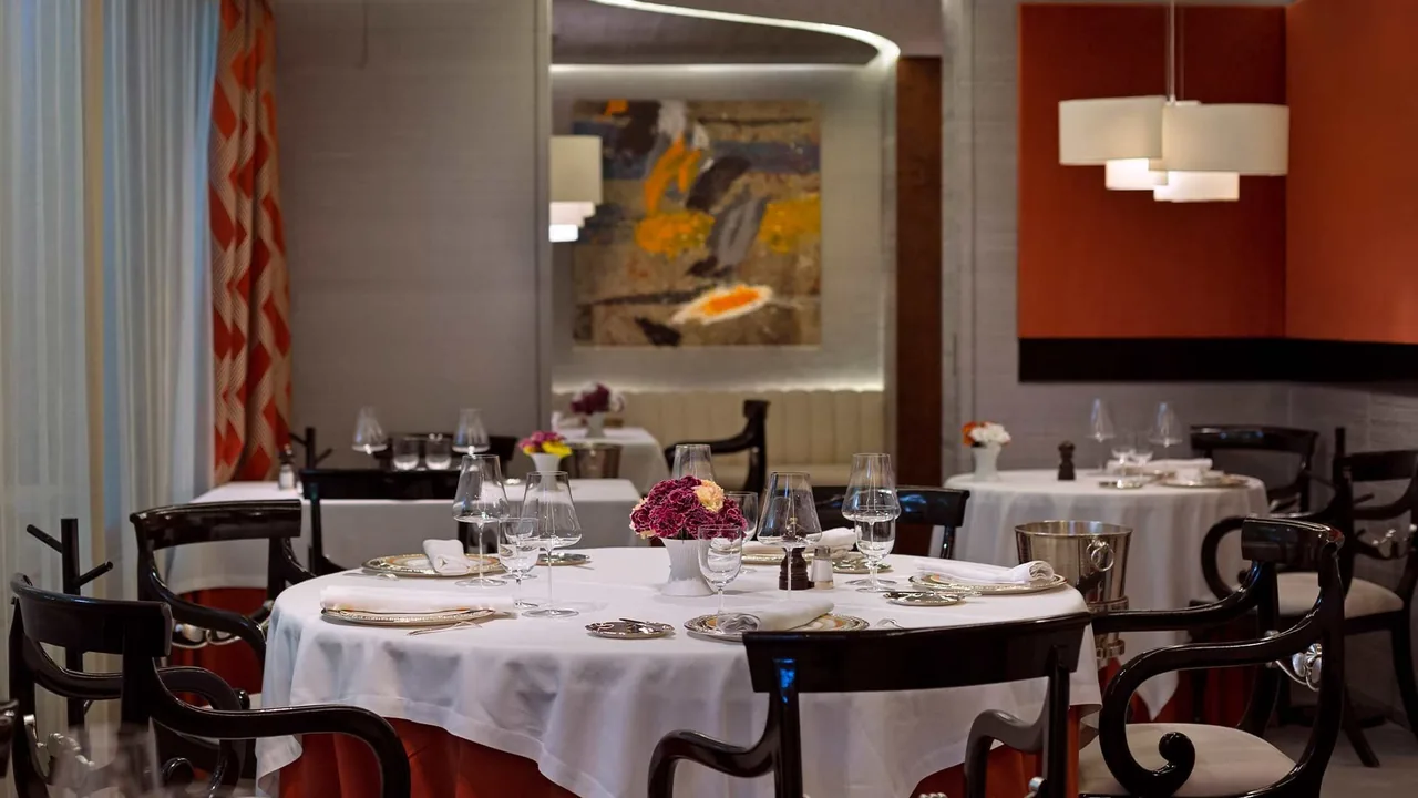A dining room with round tables set with white tablecloths, dark chairs and an orange accent wall.