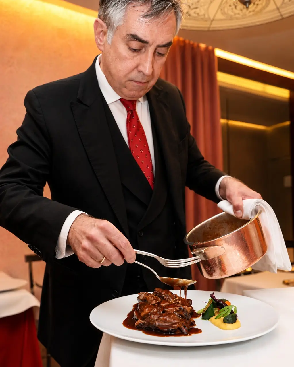 Waiter in black tie ladling dark sauce from a copper pot over a plated dish tableside, with purée and greens