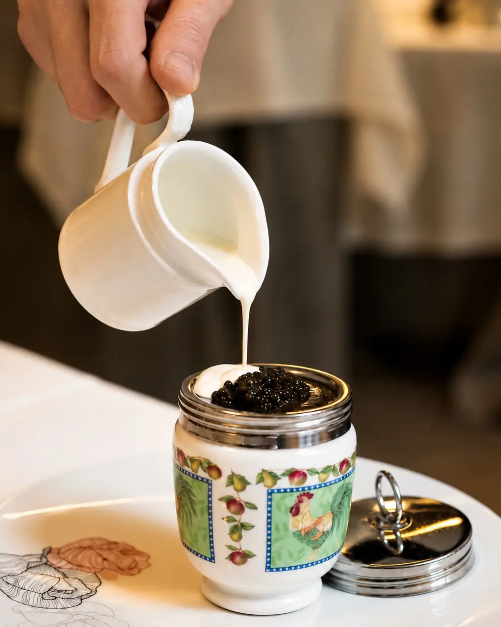 Hand pouring cream from a small jug onto caviar in a porcelain egg-shaped server
