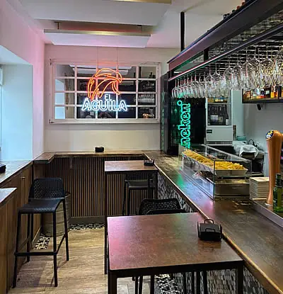 Bar area with wooden counter, black stools, hanging wine glasses and orange 'El Aguila' and green 'Heineken' neon signs